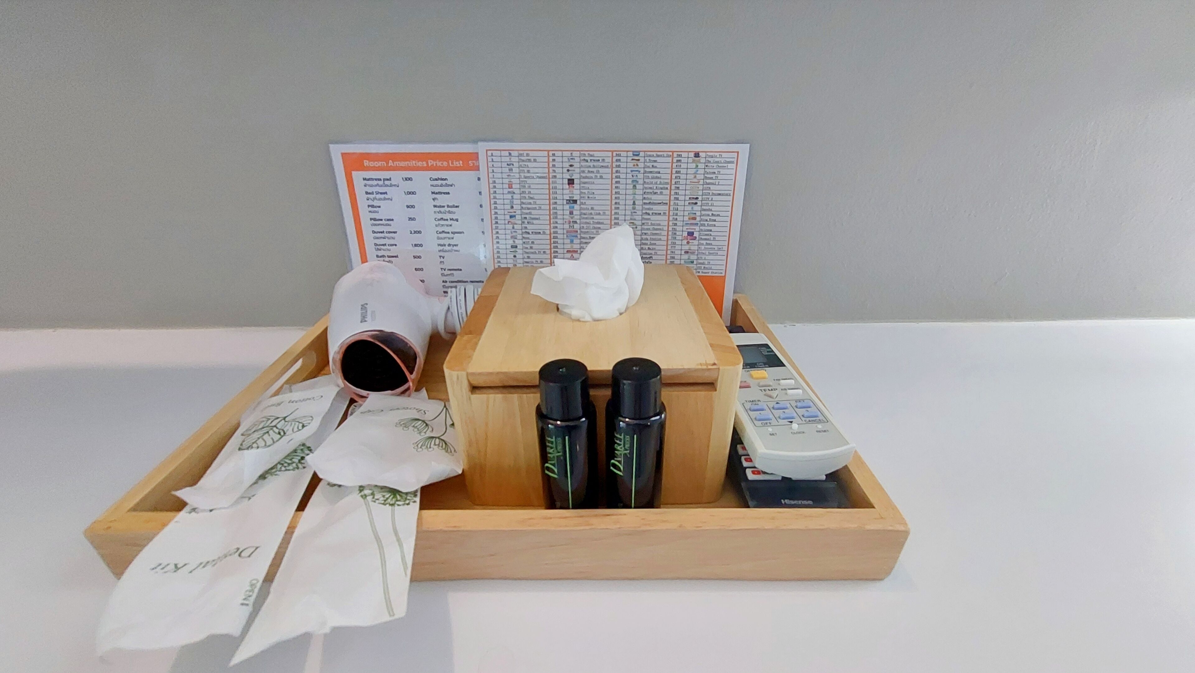 A wooden tray with toiletries, a remote control, and a tissue box, with a room amenities price list in the background.