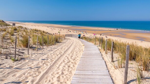 Une promenade en bois menant à une plage de sable où se trouvent des gens et des parasols.