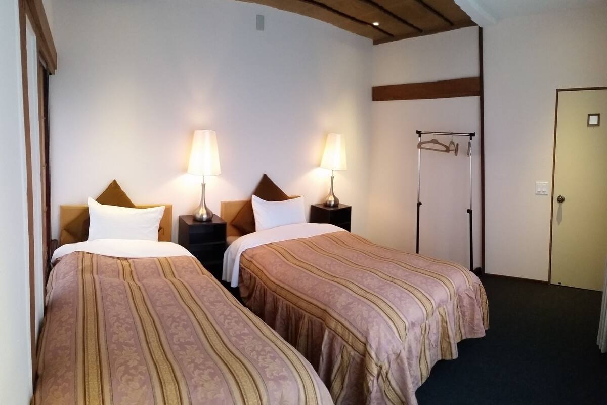 Une chambre d’hôtel avec deux lits, chacun doté d’une tête de lit et d’une table de chevet avec une lampe. On peut voir un porte-manteau et une porte dans la pièce.