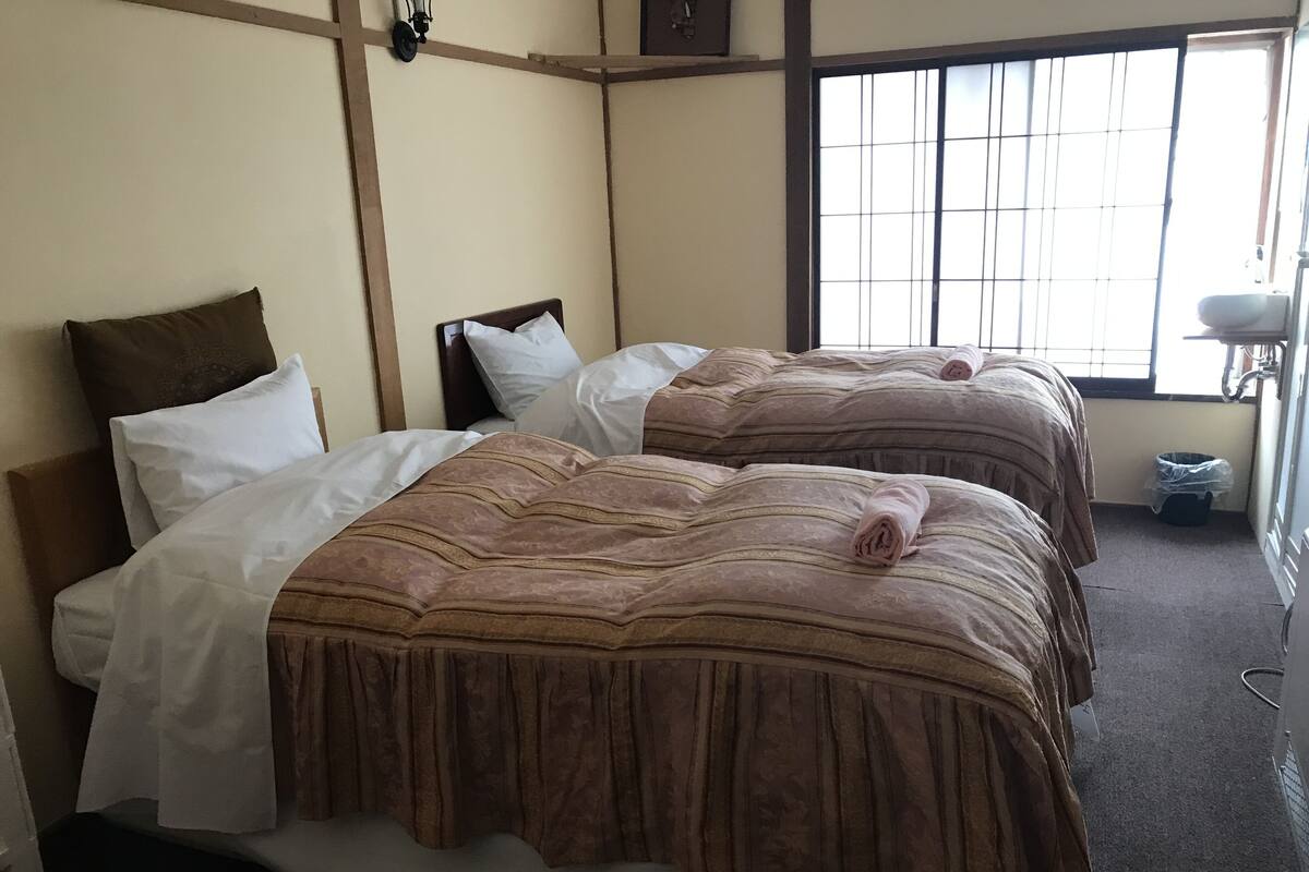 Une chambre d’hôtel avec deux lits, chacun recouvert d’une couverture marron et agrémenté d’oreillers blancs. On y trouve une fenêtre avec des barreaux, une petite table avec une lampe et une poubelle.