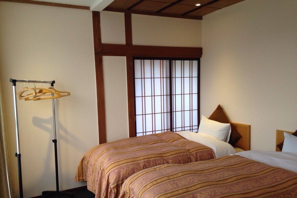 Une chambre d’hôtel avec deux lits, une fenêtre avec des stores, une tête de lit en bois et un porte-vêtements.