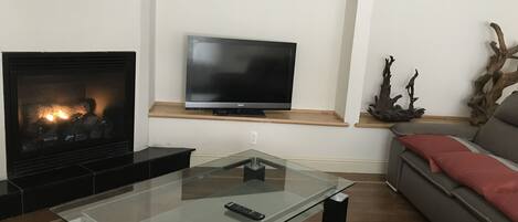Una sala de estar con chimenea, un televisor de pantalla plana sobre una estantería de madera, una mesa de centro de vidrio con un control remoto y un sofá gris con cojines rojos.