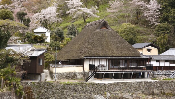 A traditional Japanese house with a thatched roof, surrounded by cherry blossoms and a stone wall. - Kanau (Gojo)