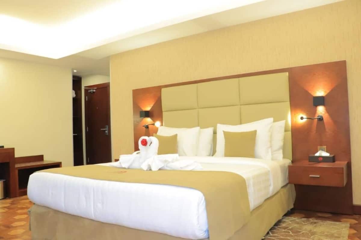 A hotel room with a large bed, a wooden nightstand, a bedside table, and a wall-mounted lamp.