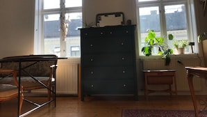 A room with a wooden dresser, a small table, and a chair. There are two windows with blinds, and a radiator is visible on the wall. - StayPlus Apartment near Vigeland Park (Oslo)