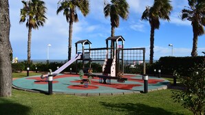 A playground with a slide, climbing structures, and swings, surrounded by palm trees and a clear sky. - Anacasa Penthouse Azul Marino Almadrava AT2202 (Denia)