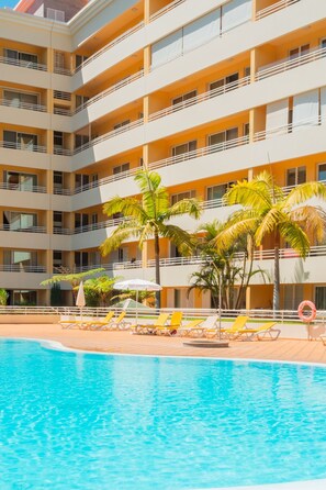 Un hotel de varios pisos con piscina, palmeras y tumbonas.