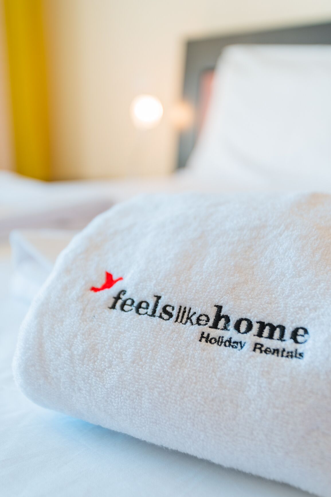 Un asciugamano bianco con il testo 'feels like home Holiday Rentals' e un piccolo logo raffigurante un uccellino rosso.