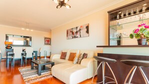 Una sala de estar moderna con un sofá blanco, una mesa de centro y un comedor con sillas y una mesa. Hay una zona de bar con taburetes y una estantería para vinos.