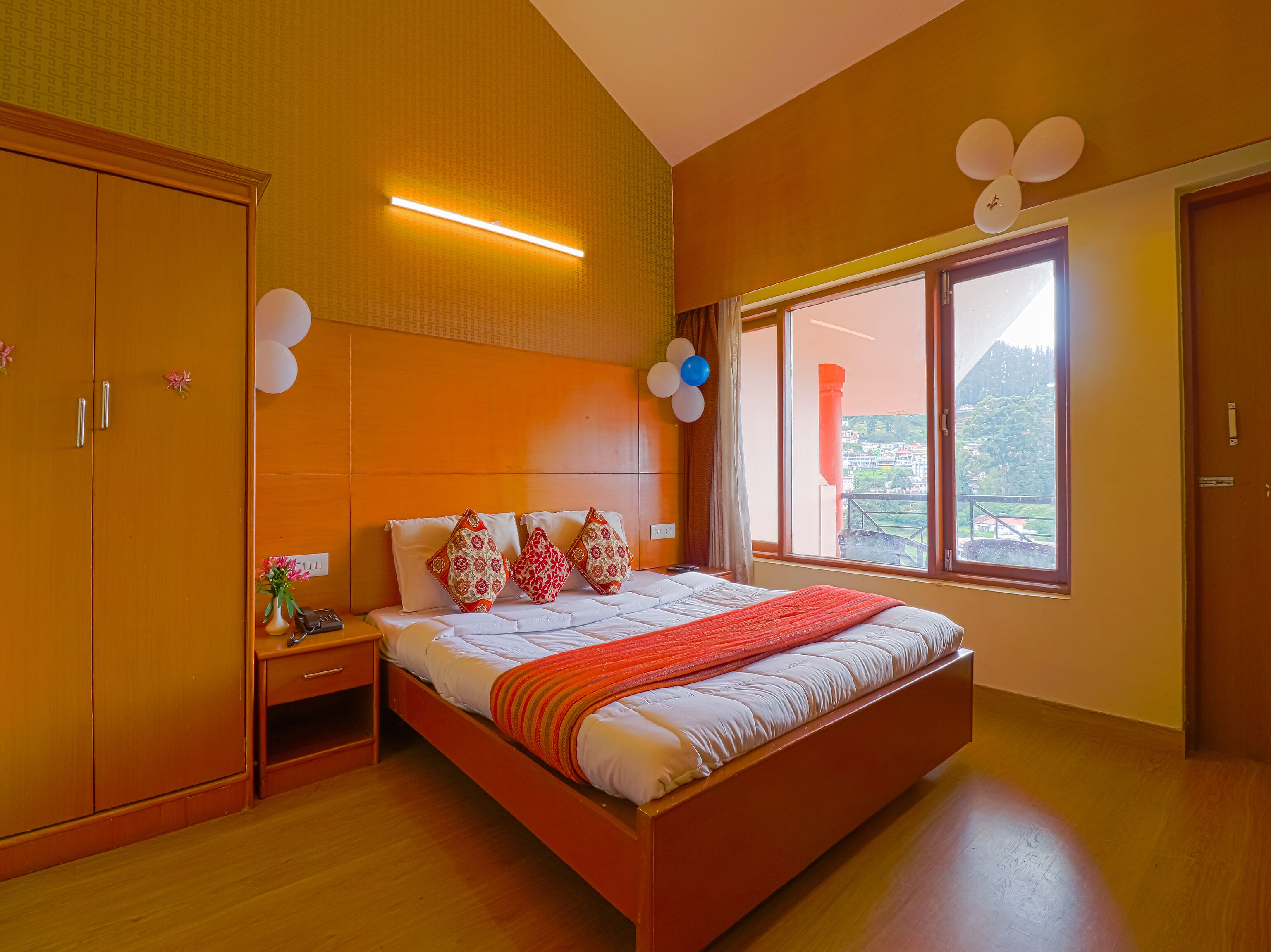 A bedroom with a large bed, wooden wardrobe, bedside table, and a window with a view of greenery.