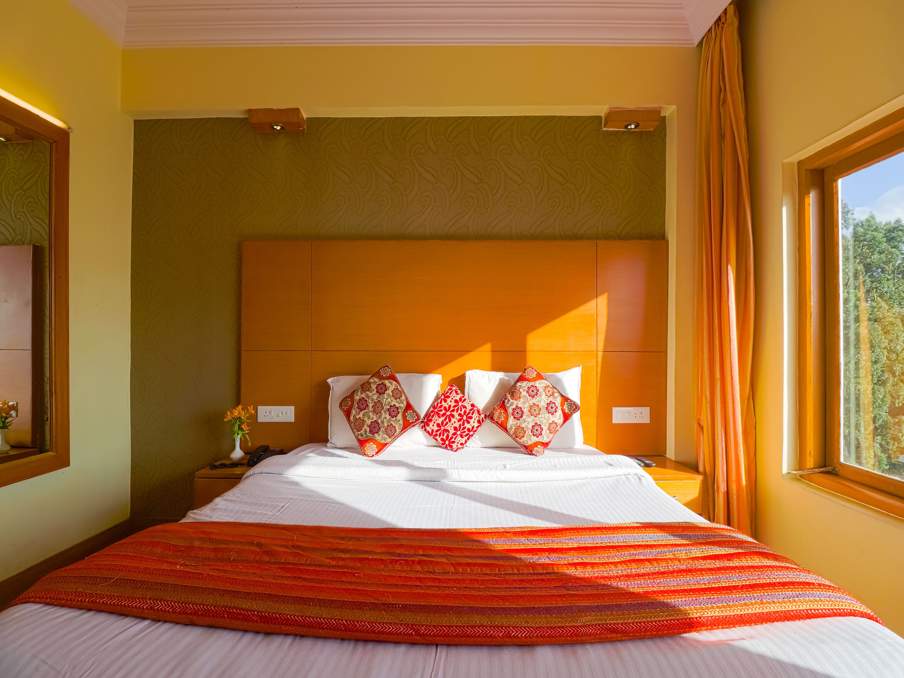 Quarto de hotel com uma cama grande, uma mesa de cabeceira, um espelho e uma janela com cortinas.