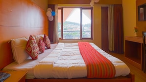 A hotel room with a large bed, a desk, a chair, a window with a view of a town, and a ceiling fan.
