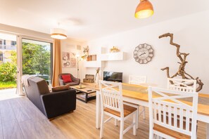 A modern living room with a sofa, dining table, and chairs. There is a TV, decorative items, and a window with a view of greenery.
