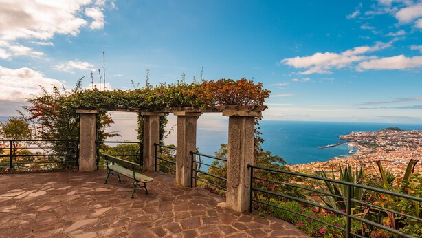 A stone-paved area with a bench and railing, overlooking a coastal city with a clear view of the sea and a distant town. - FLH Funchal Ocean View with Pool (Funchal)