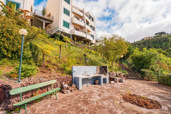 A stone-paved outdoor area with a bench, a barbecue setup, and a staircase leading to a building with balconies. - FLH Funchal Ocean View with Pool (Funchal)