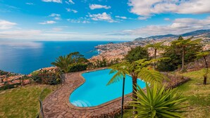 A swimming pool with a stone border, surrounded by grass and palm trees, overlooking a coastal cityscape. - FLH Funchal Ocean View with Pool (Funchal)