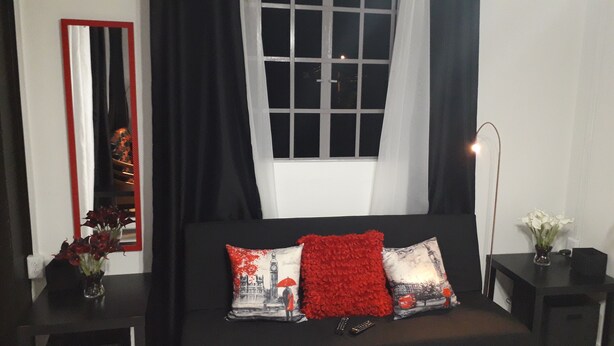 A bedroom with a black sofa, a mirror, a window with black curtains, and a bedside table with a vase of flowers. - Close to Carnival &  POS activities! Secure 1 bdr apt, mins from Port of Spain (St Anns)
