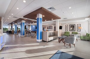 A spacious hotel lobby with a high ceiling, multiple seating areas, and a large window providing a view of the outdoors.