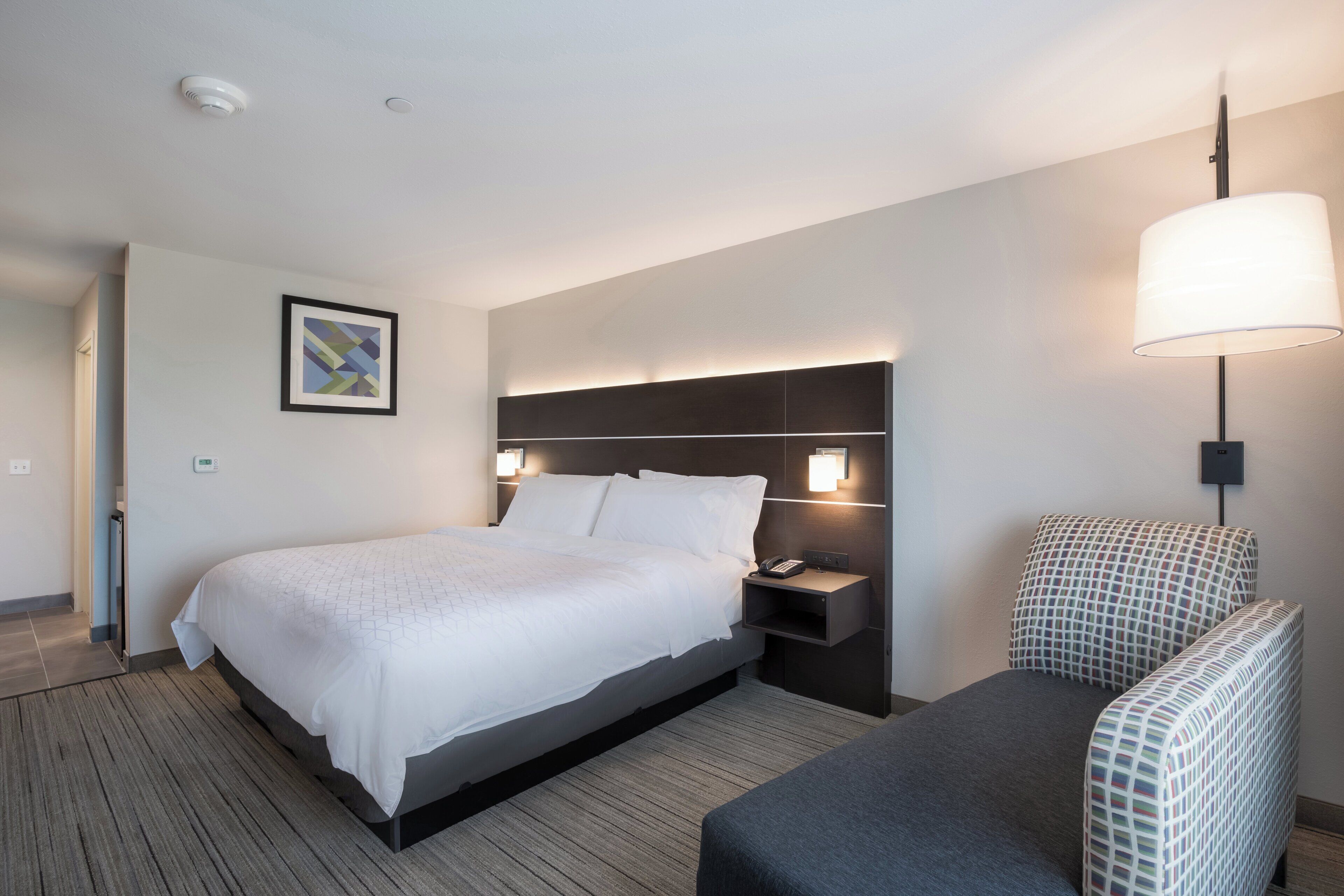 A hotel room with a bed, bedside table, lamp, and a chair.