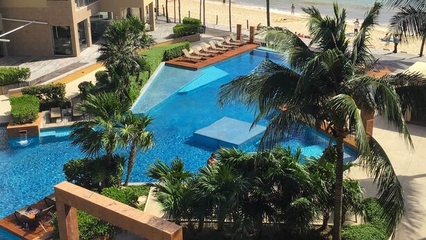 A swimming pool with a waterfall feature, surrounded by palm trees and lounge chairs, with a beach in the background. - Playa del Carmen Beachfront Condo w/ Pool Access! (Playa Del Carmen)