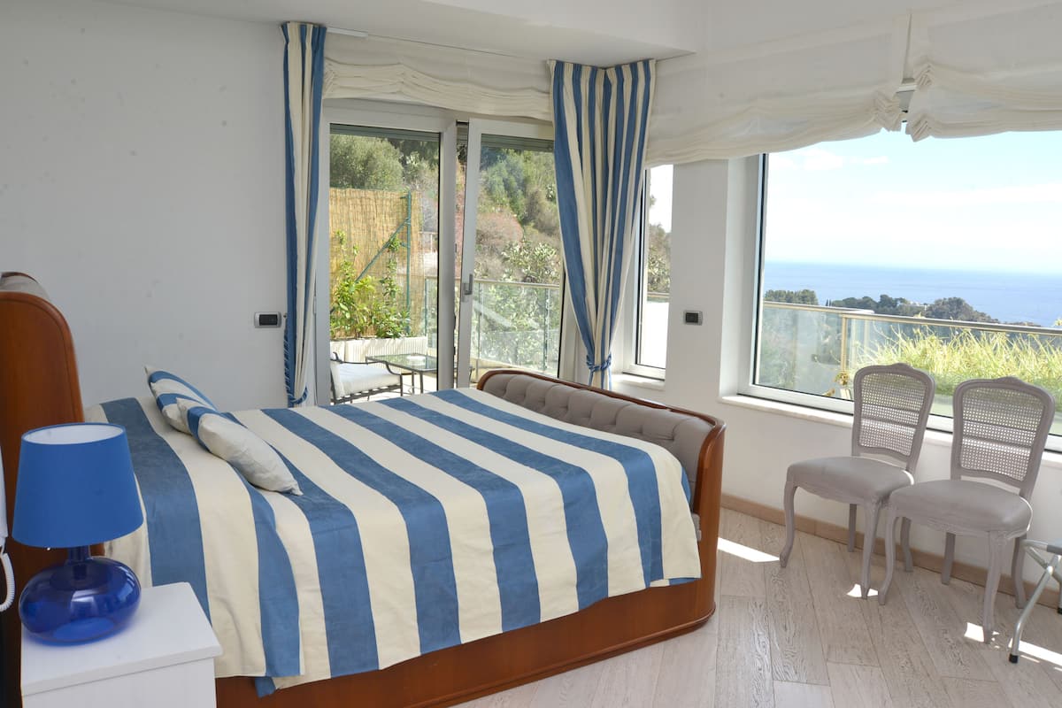 Ein Schlafzimmer mit Bett, Nachttisch, Lampe, zwei Stühlen und einem großen Fenster mit Meerblick.