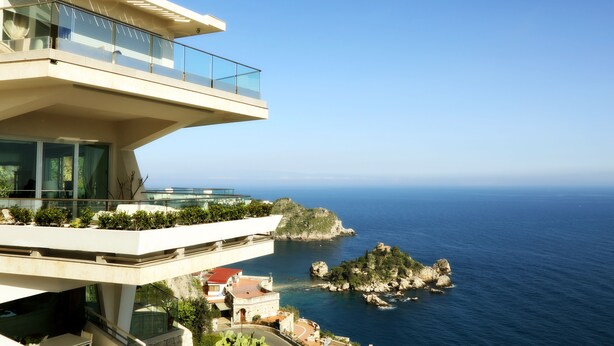 A modern building with a glass balcony overlooking a coastal area with rocky terrain and the sea. - Maison Blanche Luxury B&B Taormina (Taormina)