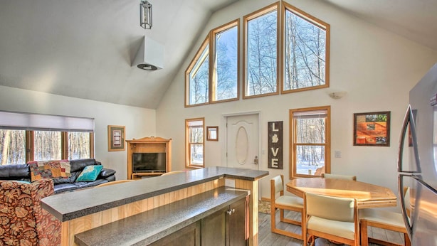 A modern kitchen with a central island, wooden dining table, and chairs. There is a built-in oven and a refrigerator on the right. The kitchen has large windows and a view of trees outside. - Lakes of the North Home on Snowmobile & ATV Trail! (Mancelona)