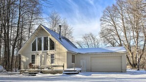 A two-story house with a garage, a covered porch, and a snow-covered roof. - Lakes of the North Home on Snowmobile & ATV Trail! (Mancelona)