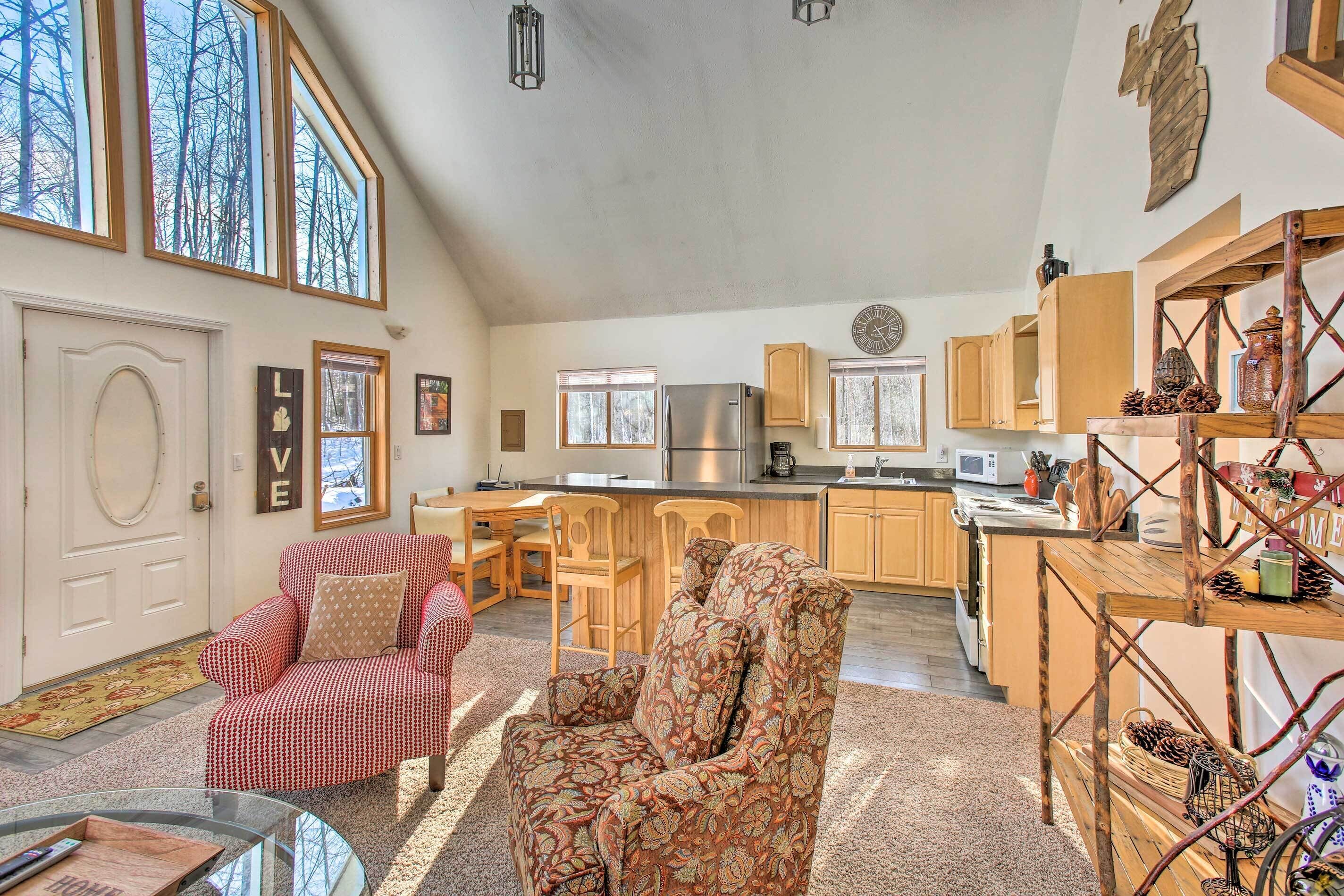 A spacious living room with a red and white patterned sofa, a patterned armchair, and a glass coffee table. The kitchen in the background features wooden cabinets and a stainless steel refrigerator.
