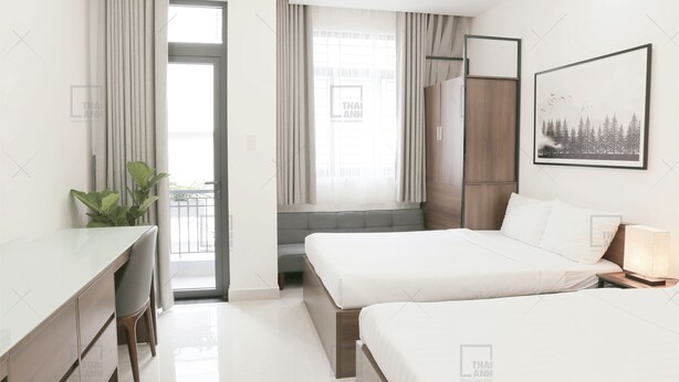 A modern hotel room with a desk, two beds, a large window with curtains, a plant, and a framed picture on the wall.