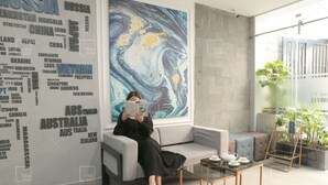A modern hotel lobby with a large abstract mural, a seating area with a sofa and a coffee table, and a glass partition with potted plants.