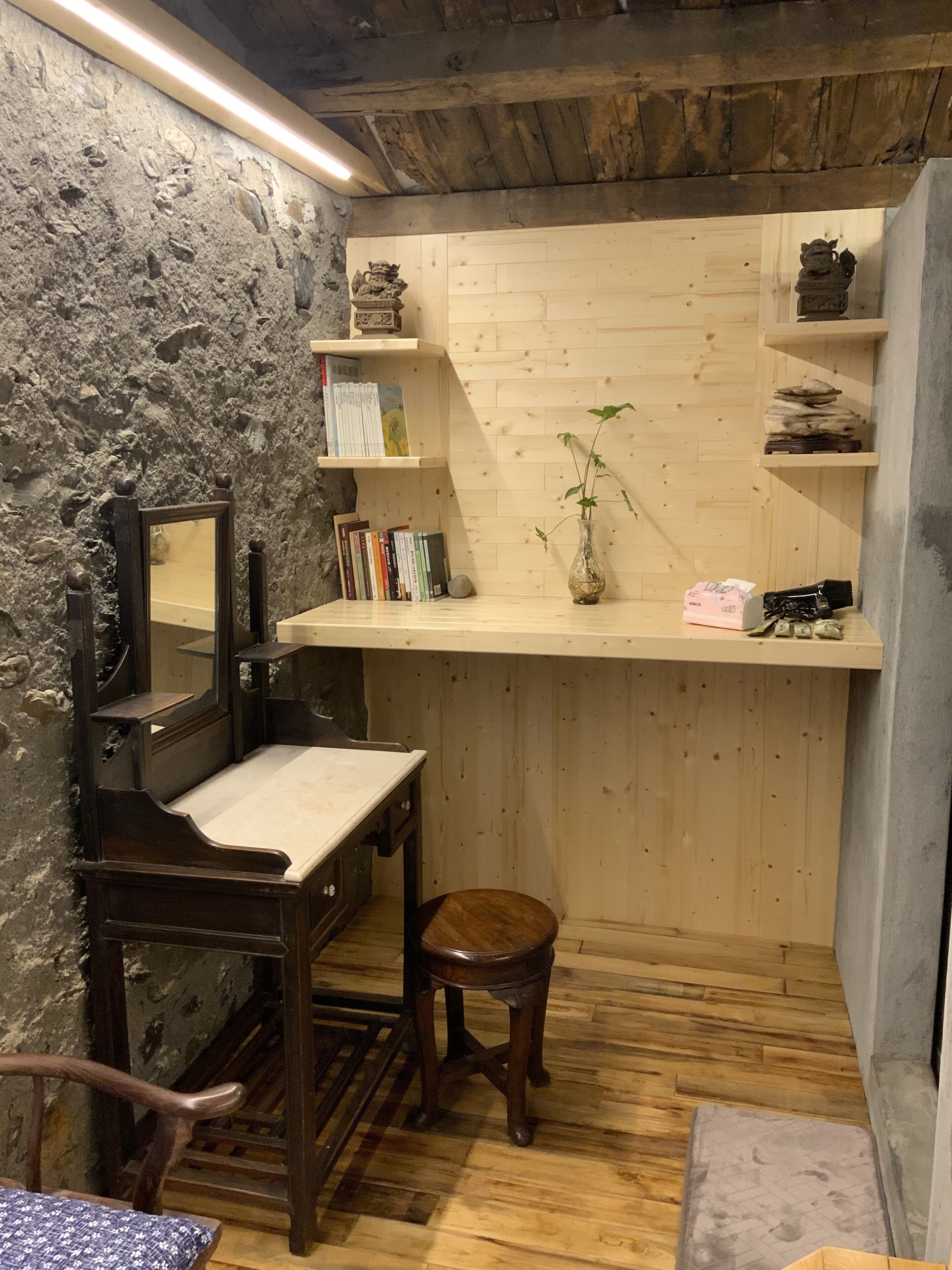 A rustic room with a stone wall, wooden shelves, a mirror, a desk, a stool, and a small plant.