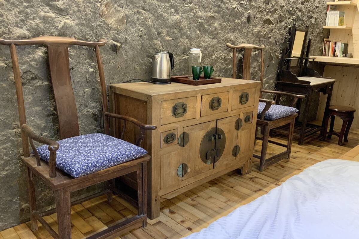 Un aparador de madera con cajones y un espejo, una silla de madera con cojín, una cama con ropa blanca, y una pared de piedra.