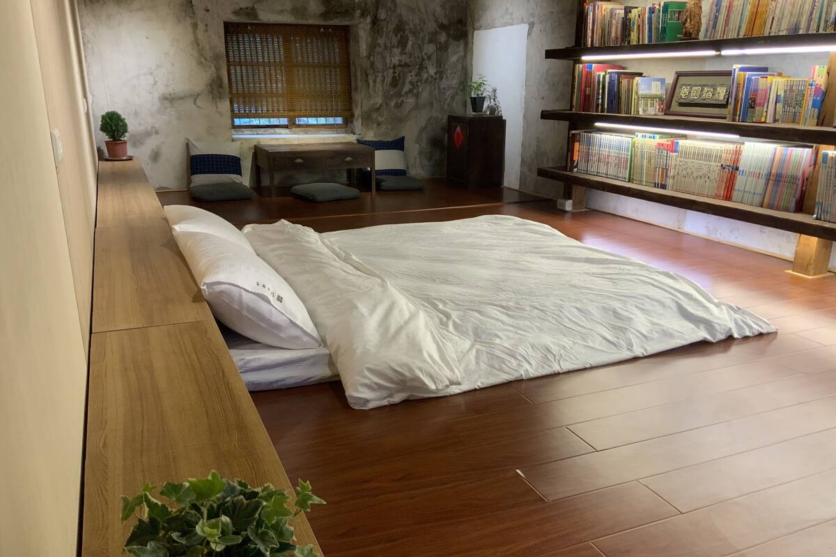 Un dormitorio con piso de madera, una cama con ropa blanca, una mesita de noche pequeña, una estantería con libros y una ventana con vistas al exterior.