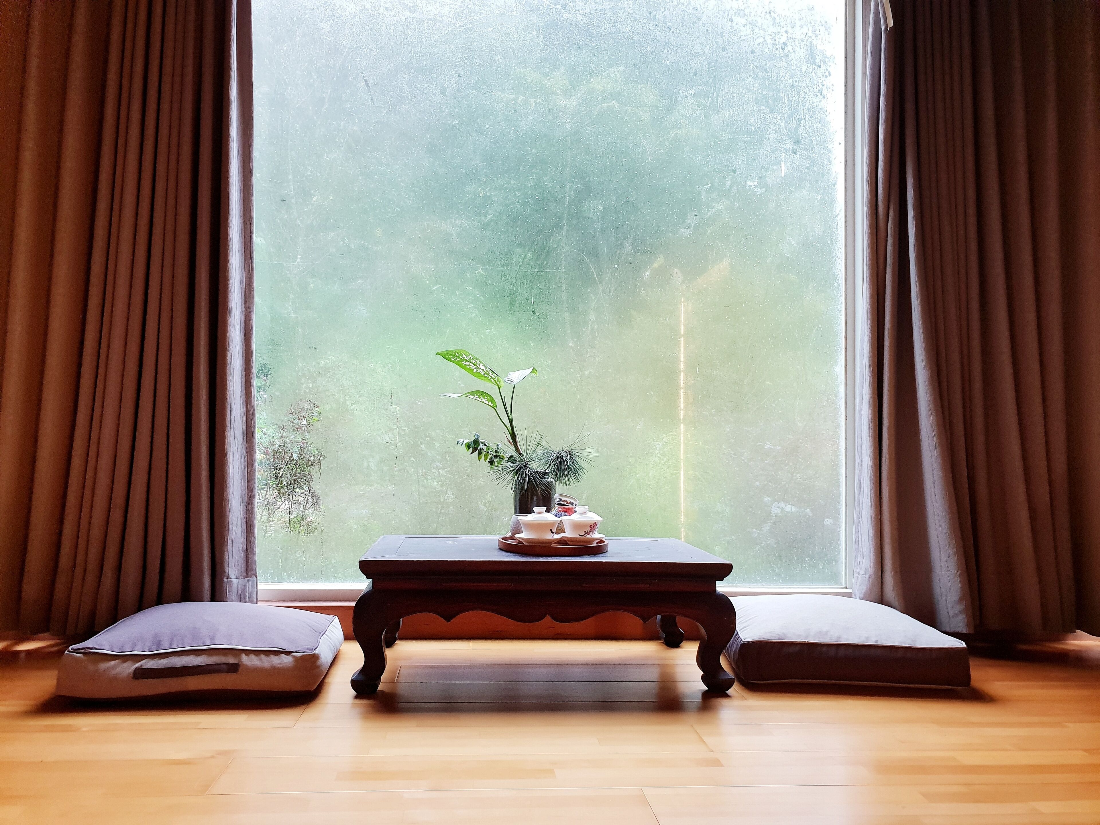 Una habitación con un ventanal grande, una mesa de madera con un florero y dos cojines, y vistas a la vegetación exterior.