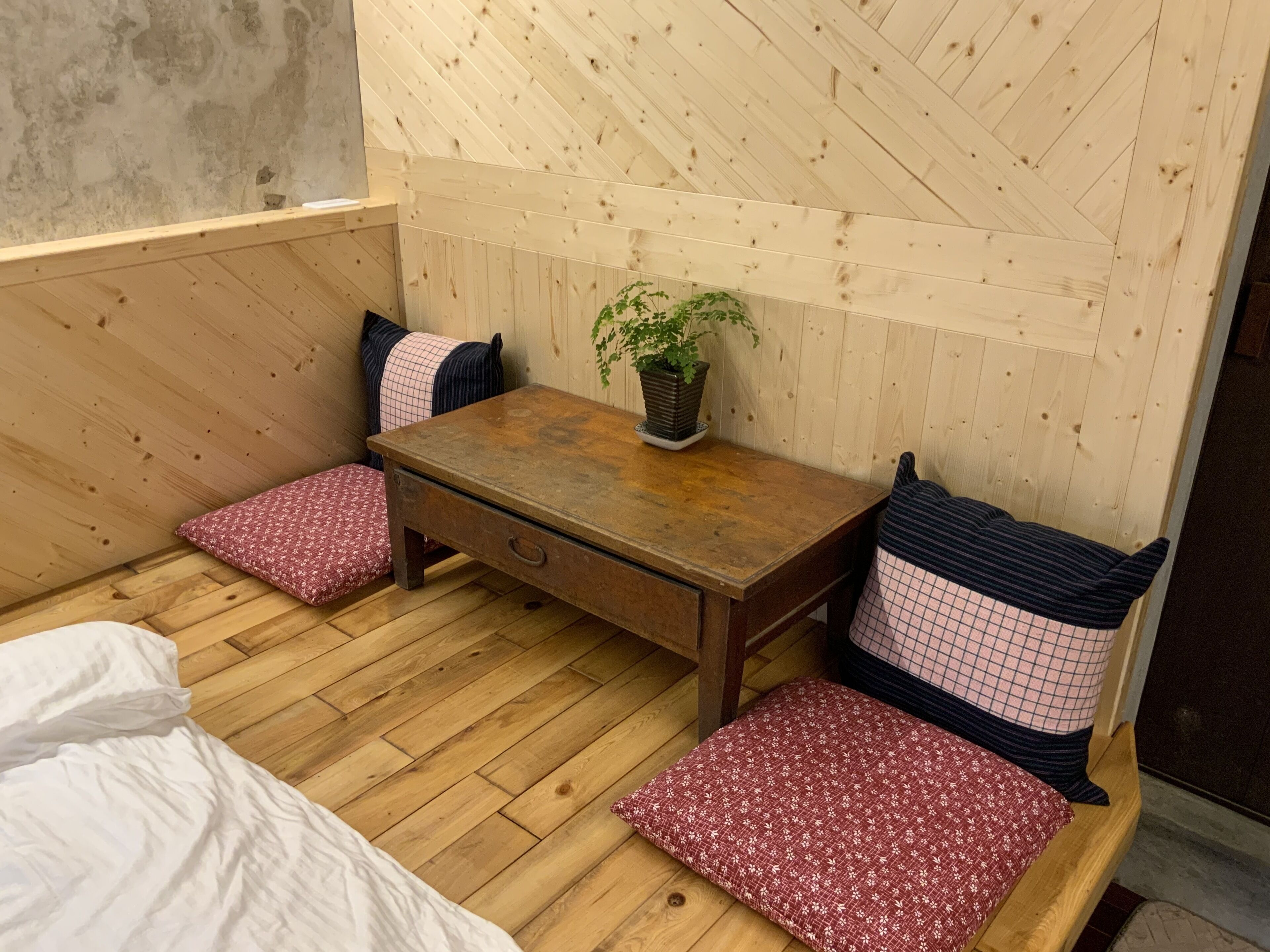 A small wooden room with a bed, a wooden table, and two chairs. There is a potted plant on the table.