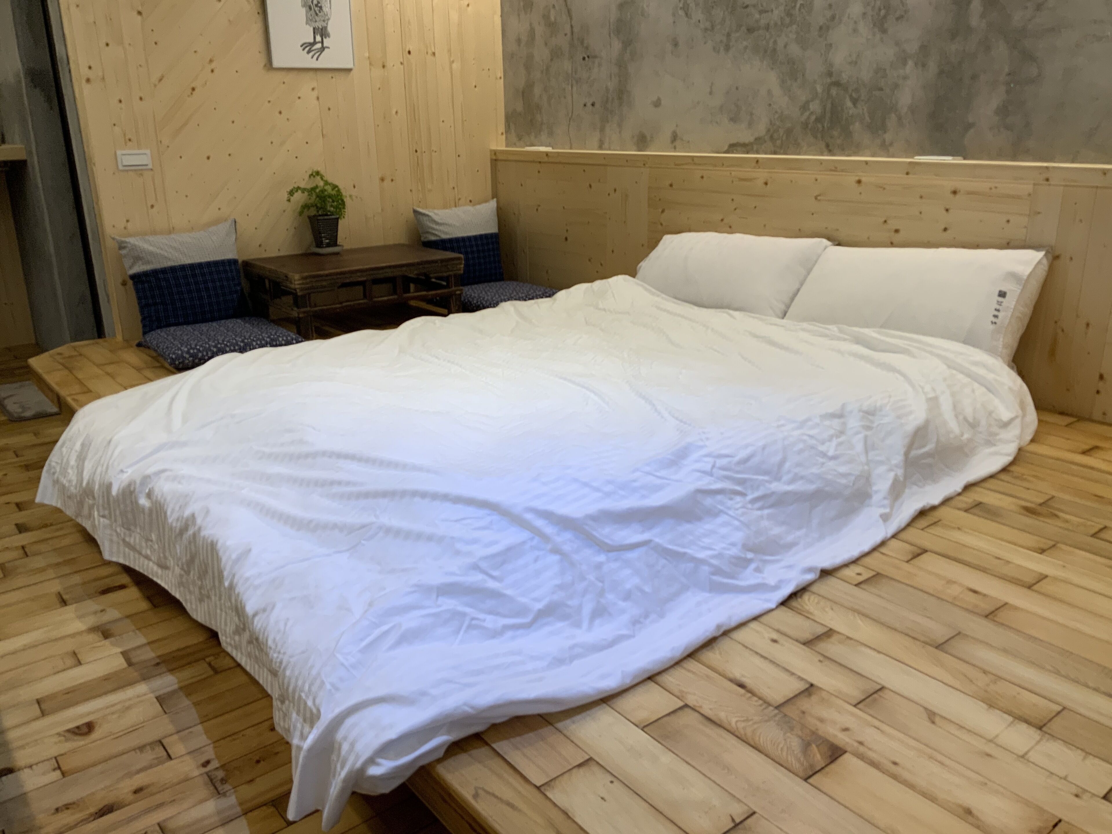 Un dormitorio con piso de madera, una cama con ropa de cama blanca, cabecera de madera y una mesita con una planta.