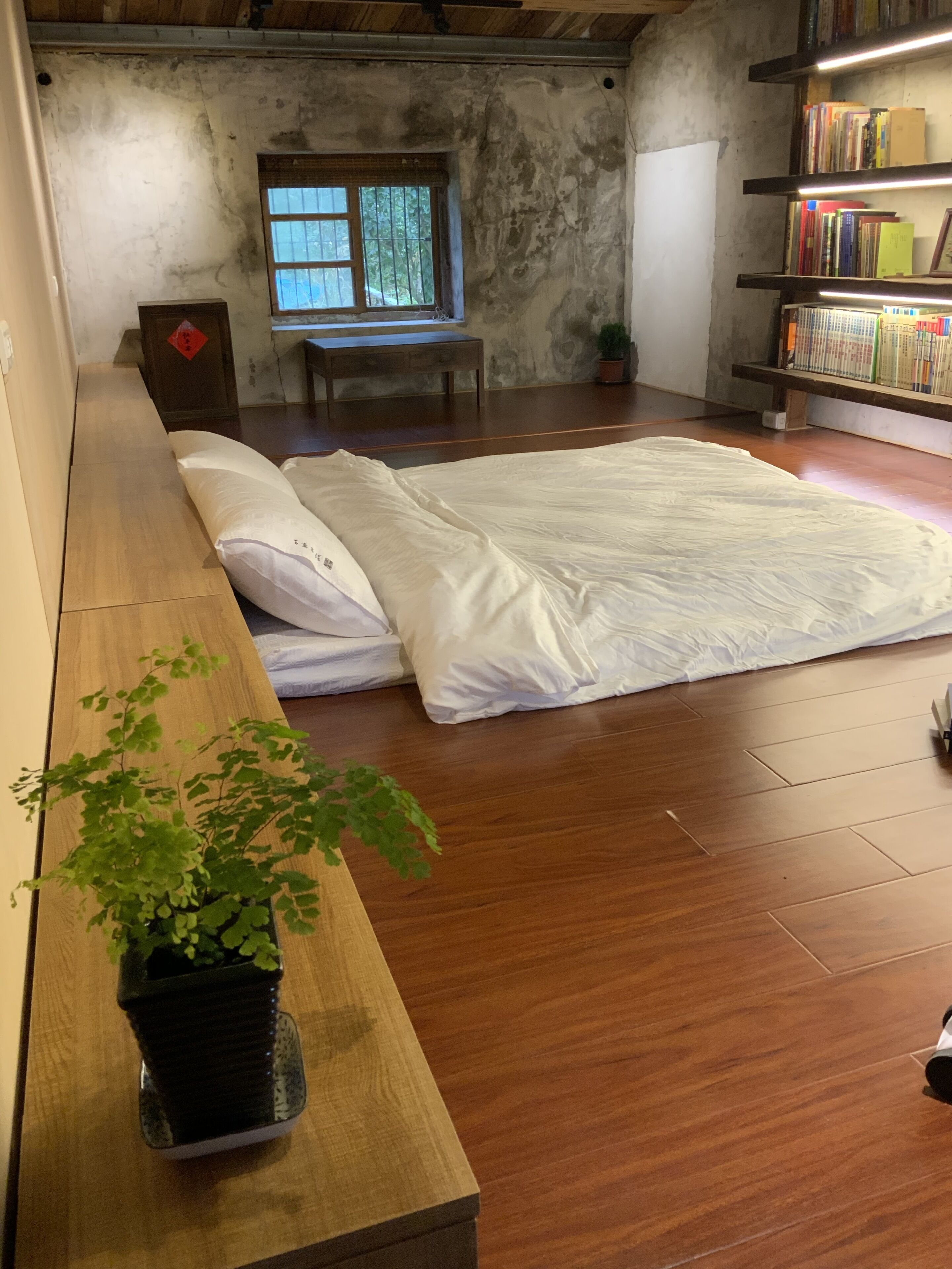A room with a large bed, wooden flooring, a small window, a bookshelf, and a potted plant.