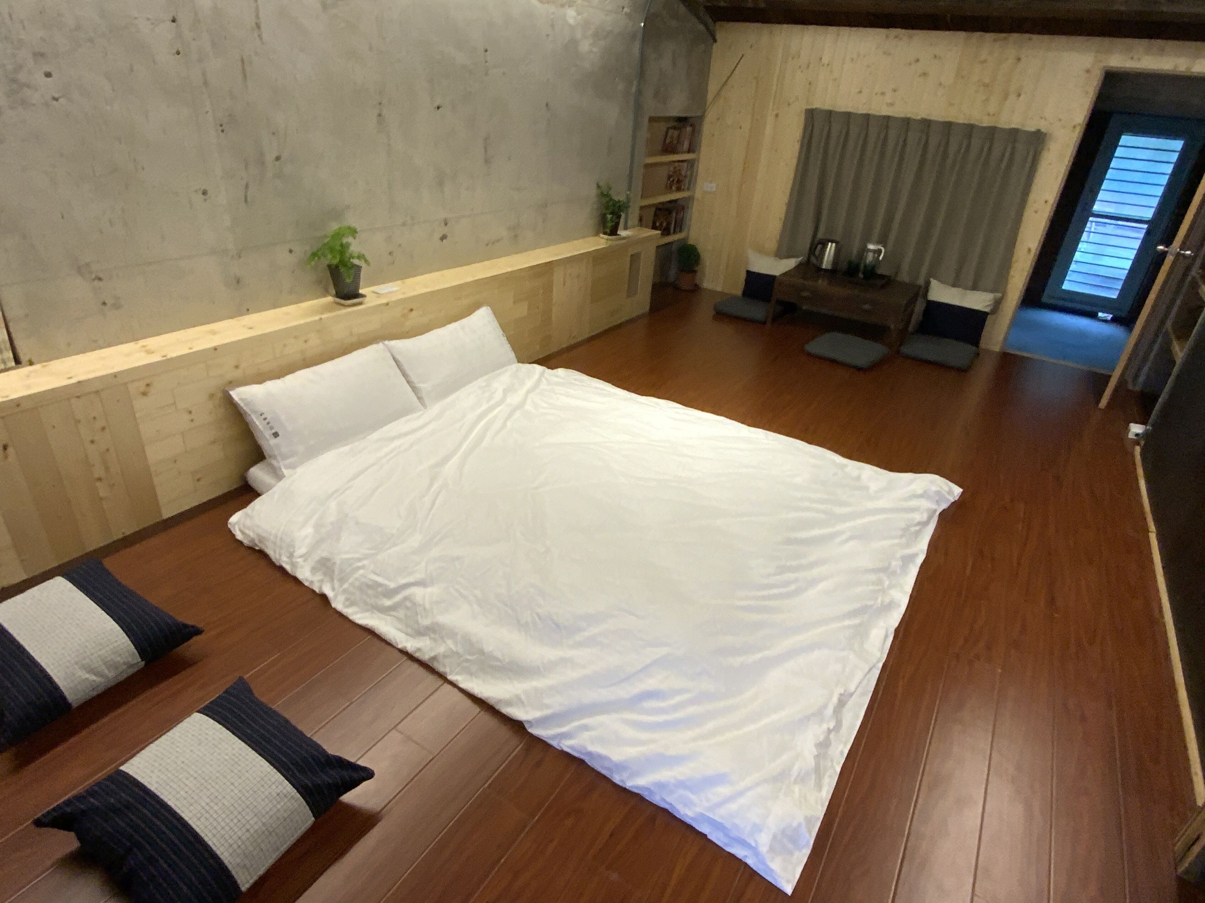 Una habitación con una cama grande, suelo de madera, pared de concreto y una pequeña zona de estar con una mesa y sillas.