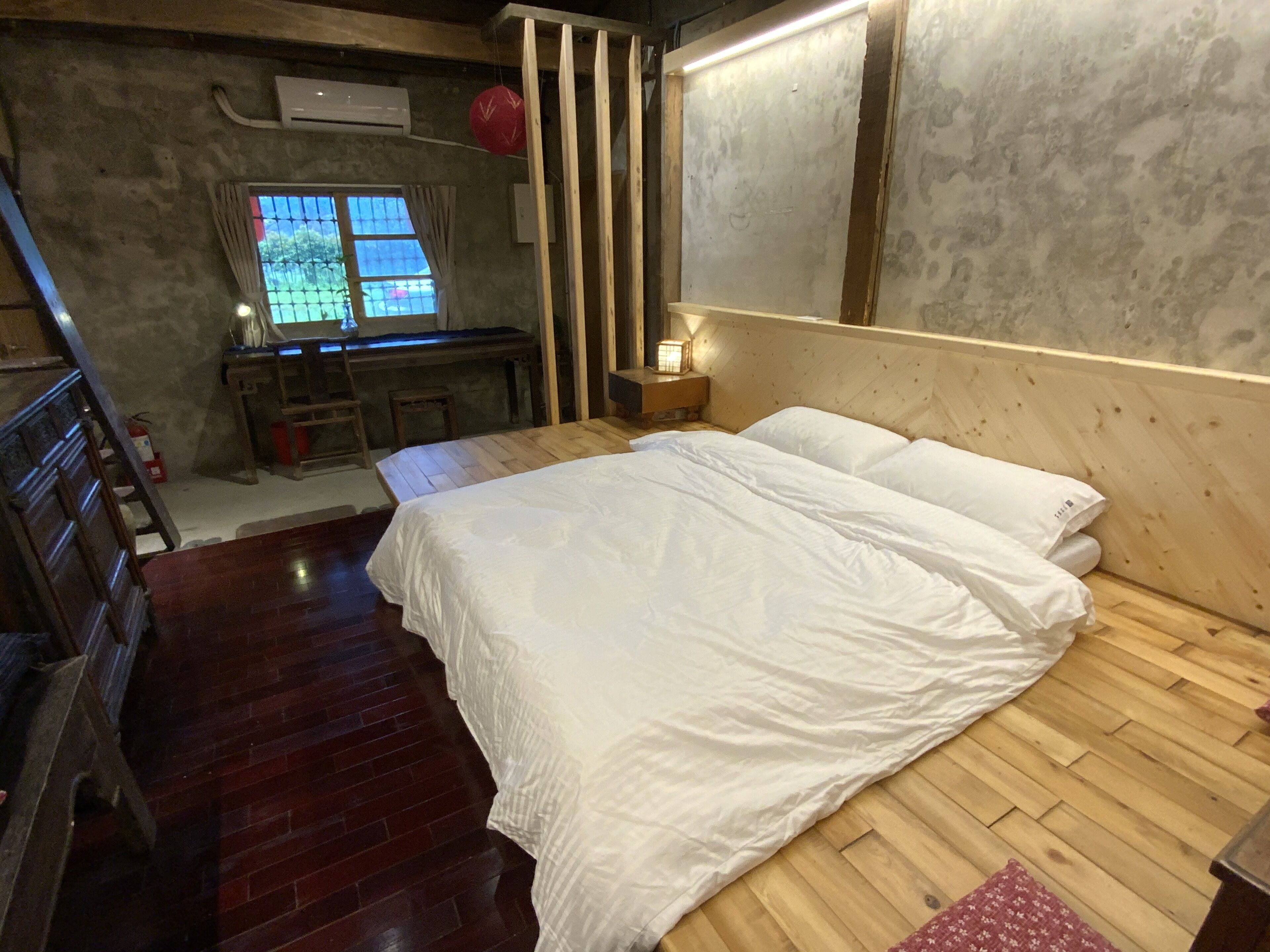 A bedroom with a wooden floor, a bed with white bedding, a nightstand with a lamp, and a window with curtains.