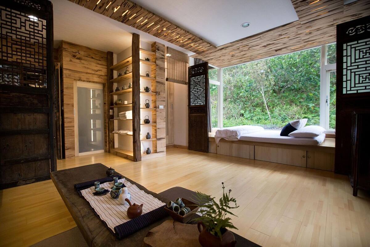 Un interior moderno con piso de madera, una zona de estar con cojines, una estantería con teteras y un ventanal con vistas a la vegetación.