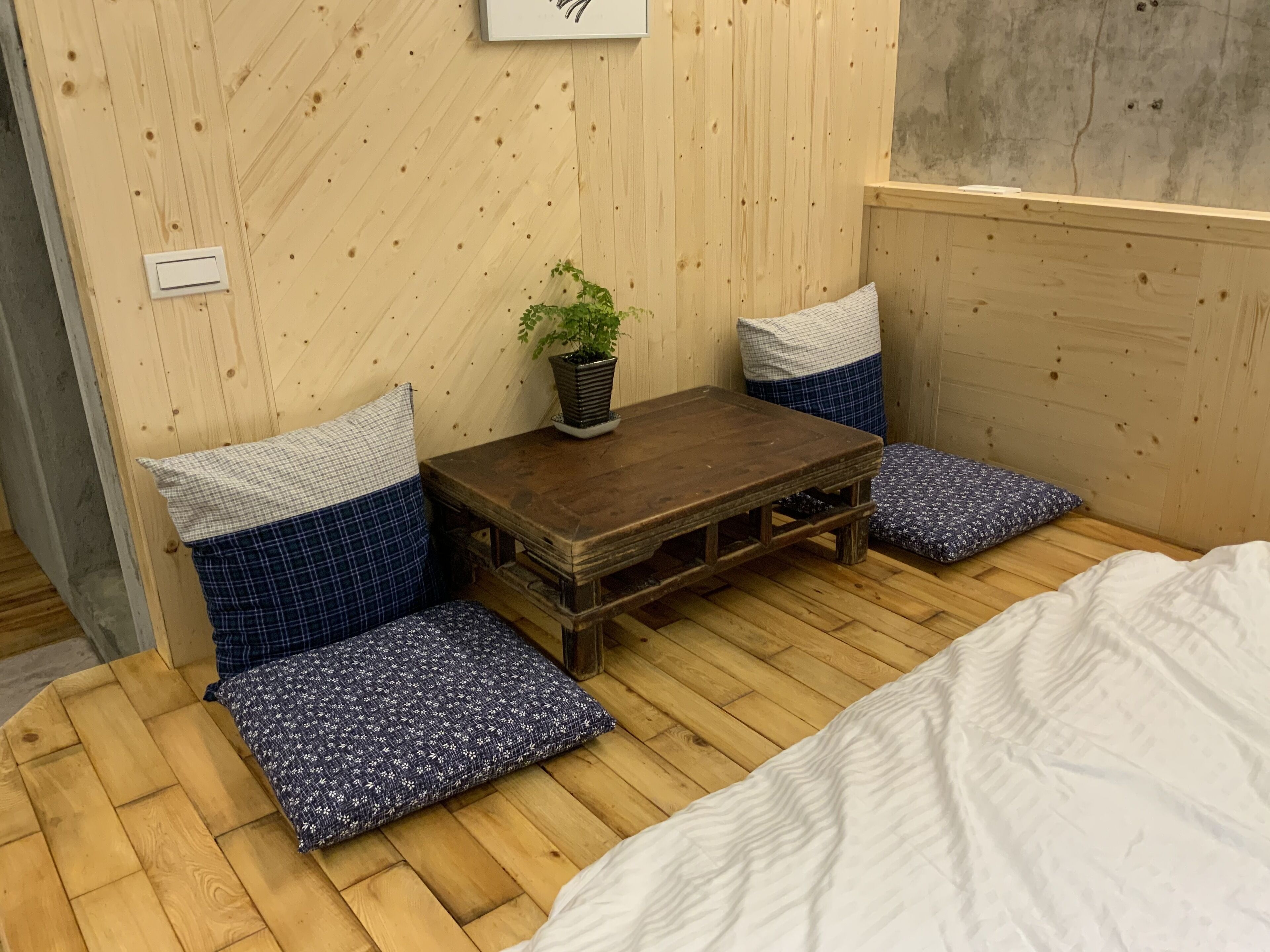 A small room with a wooden floor, a wooden bench with blue cushions, a small table with a potted plant, and a bed with white bedding.