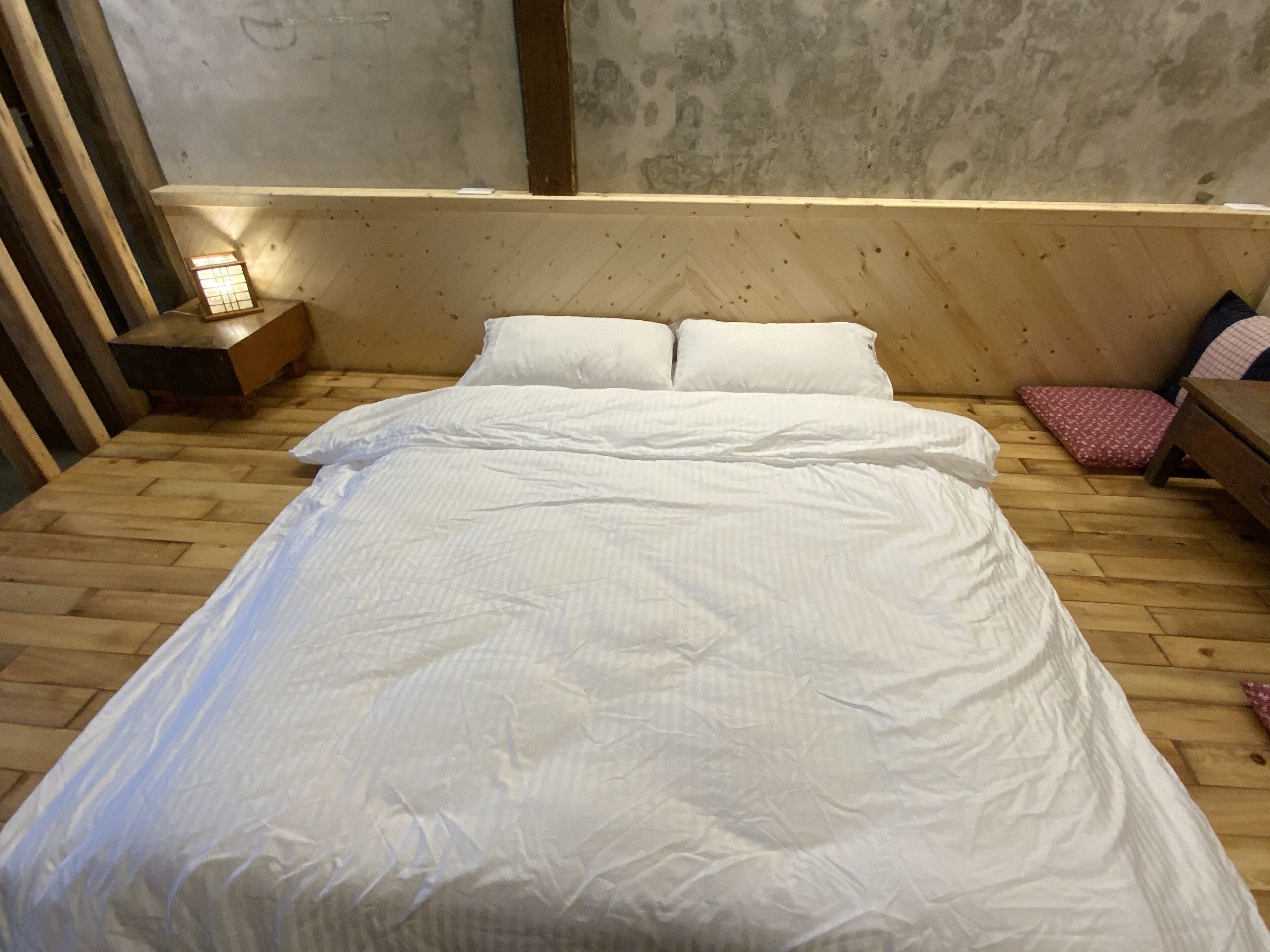 Una cama de madera con ropa de cama blanca en una habitación con pared de hormigón y suelo de madera.