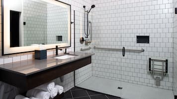 A modern bathroom with a large mirror, dual sinks, a shower with a handheld showerhead, and a hand sanitizer dispenser.
