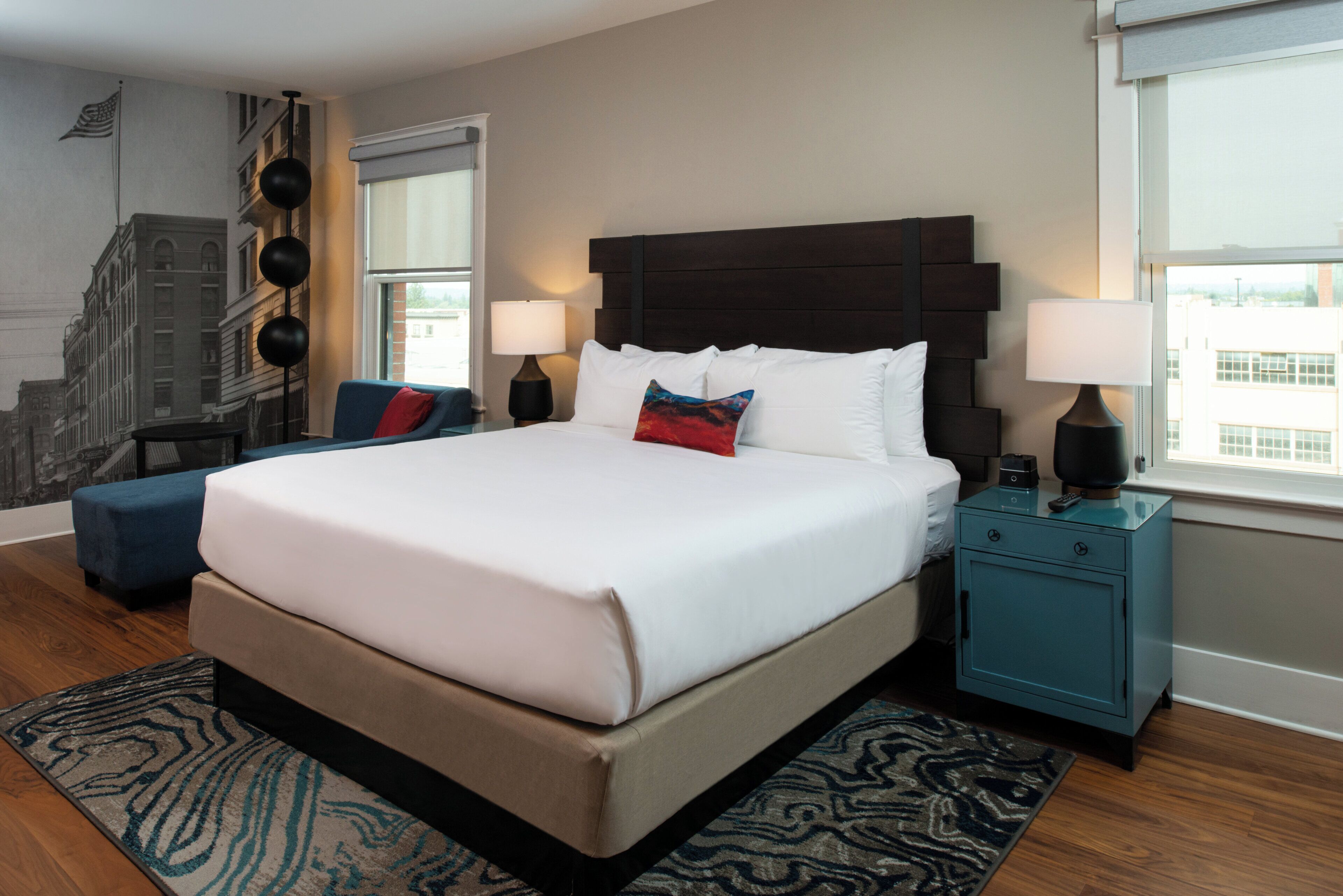 A hotel room with a large bed, two bedside tables, a blue ottoman, a patterned rug, and a cityscape mural on the wall.