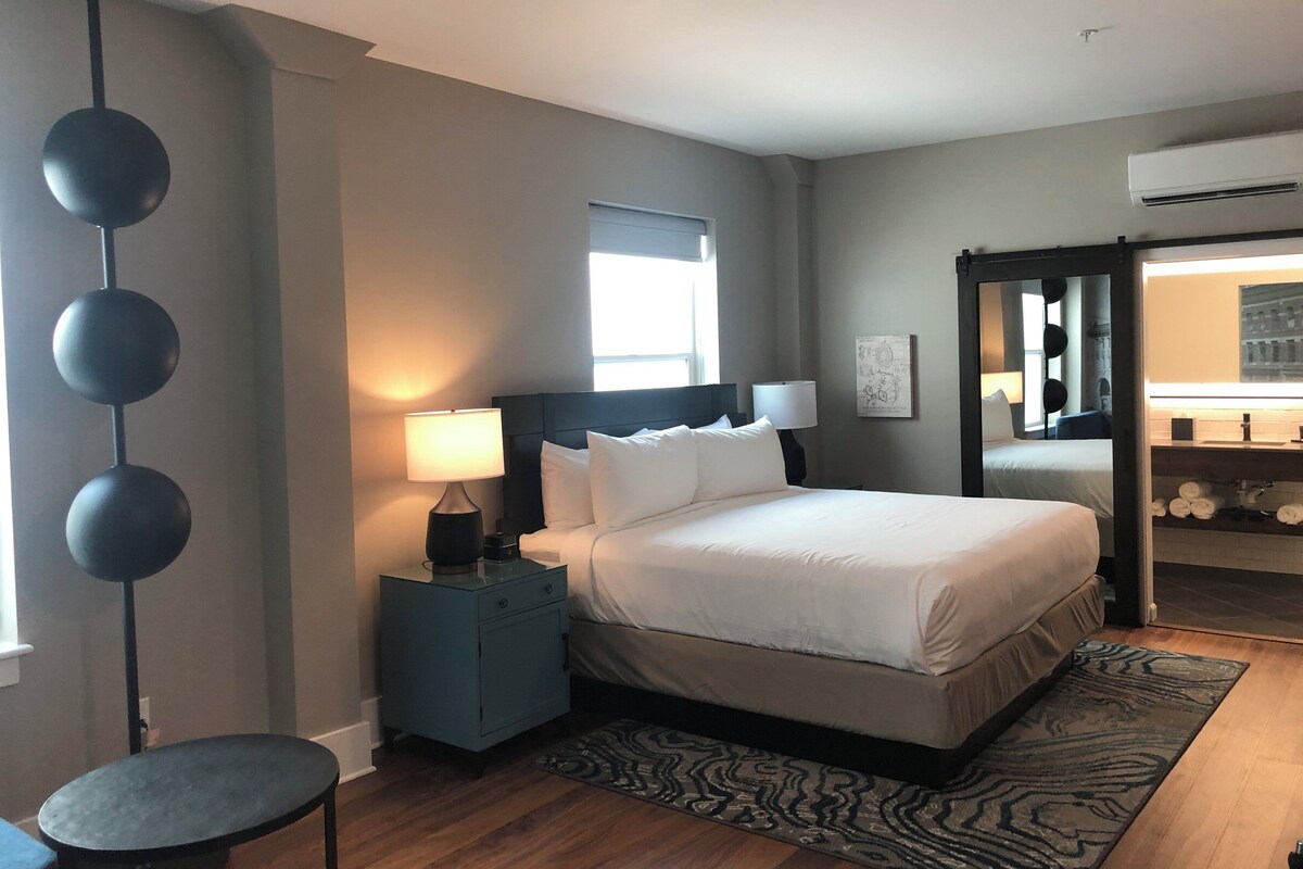 A bedroom with a bed, bedside tables, a lamp, a mirror, and a view of a bathroom.