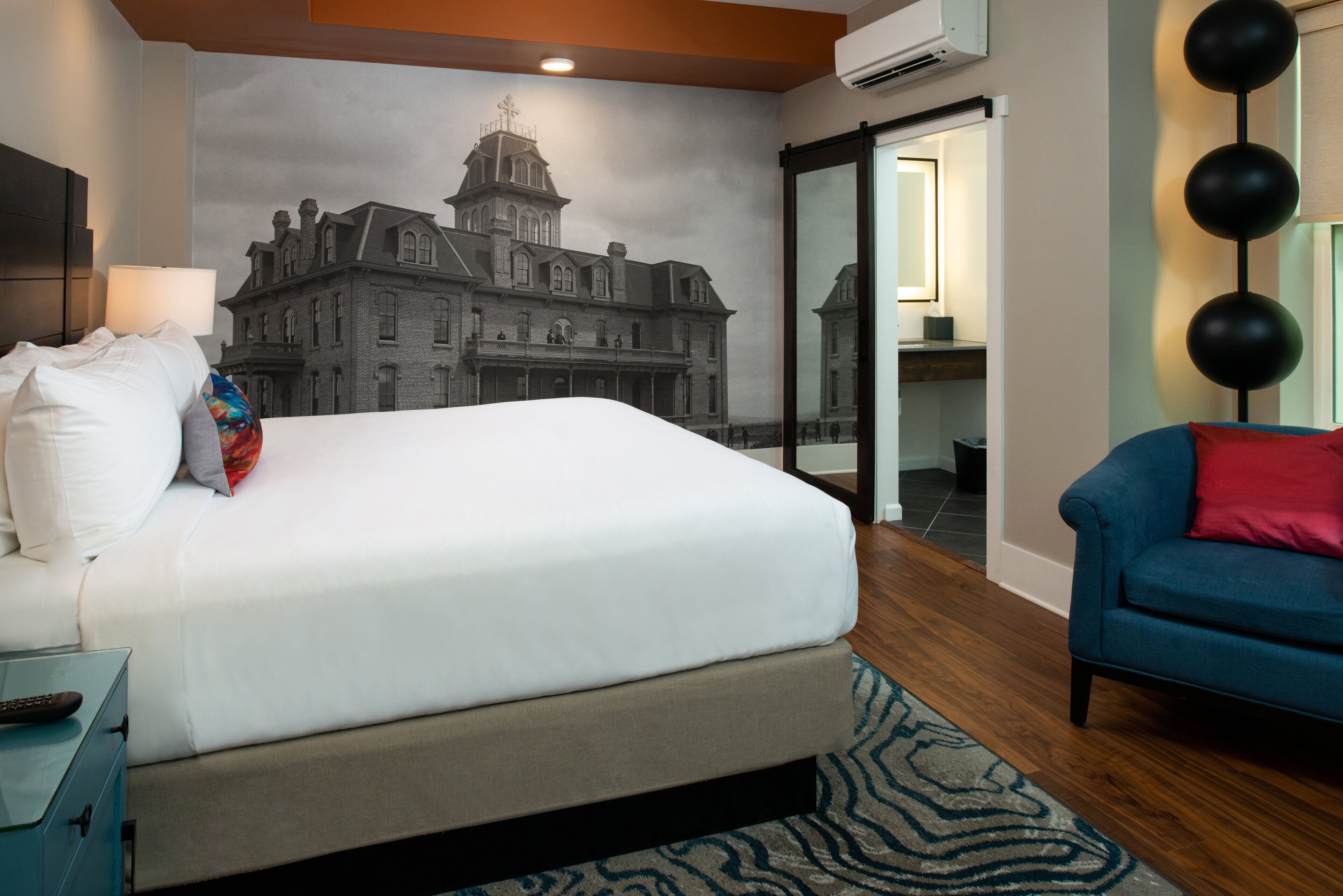 A hotel room with a large bed, a blue armchair, a wall mural, and a sliding door leading to a bathroom.