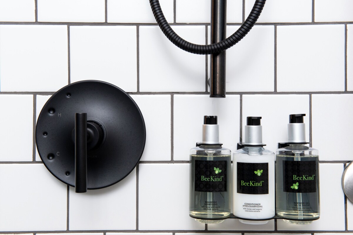 A bathroom with a black showerhead, a black circular object, and three bottles of BeeKind shower products.