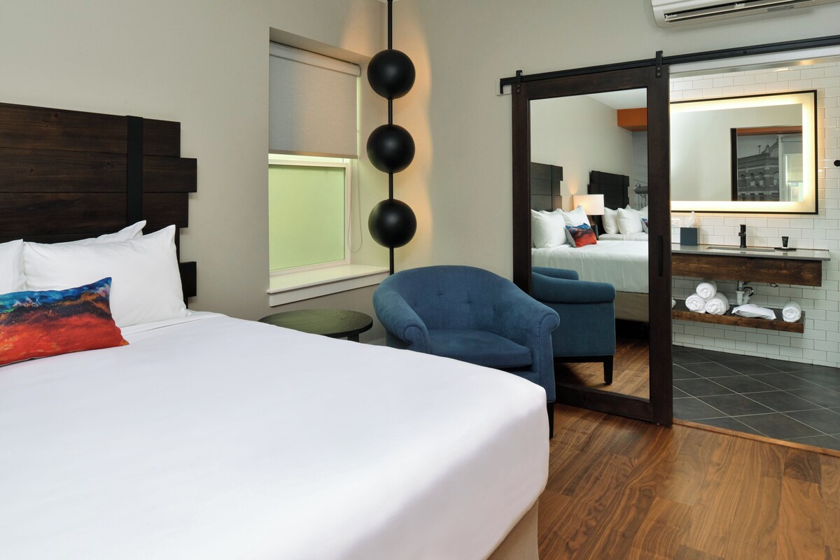 A hotel room with a bed, a blue armchair, a mirror, a window, and a bathroom with a sink and towel rack.
