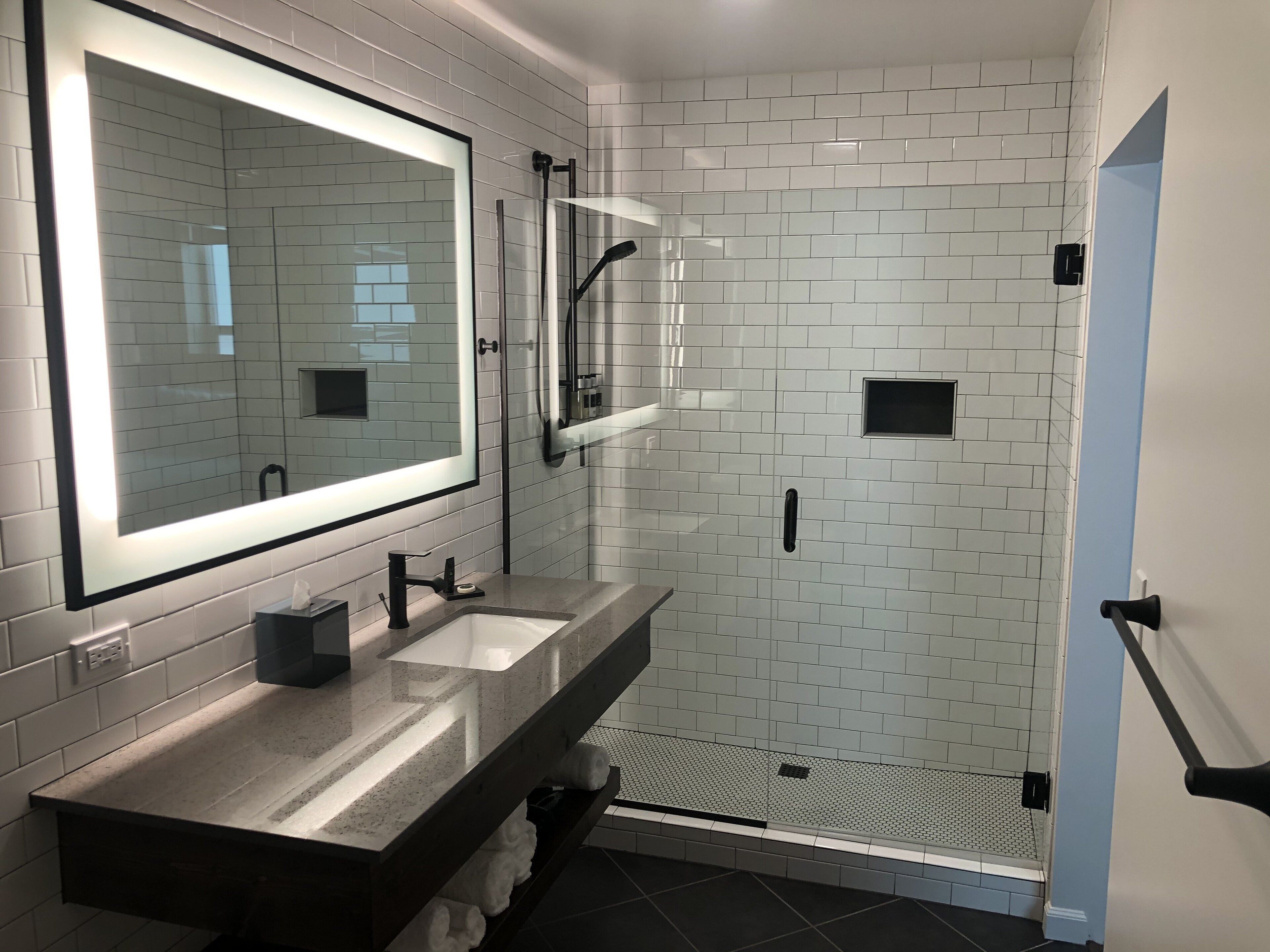 A modern bathroom with white subway tiles, a large mirror, a glass-enclosed shower, and a dark countertop.